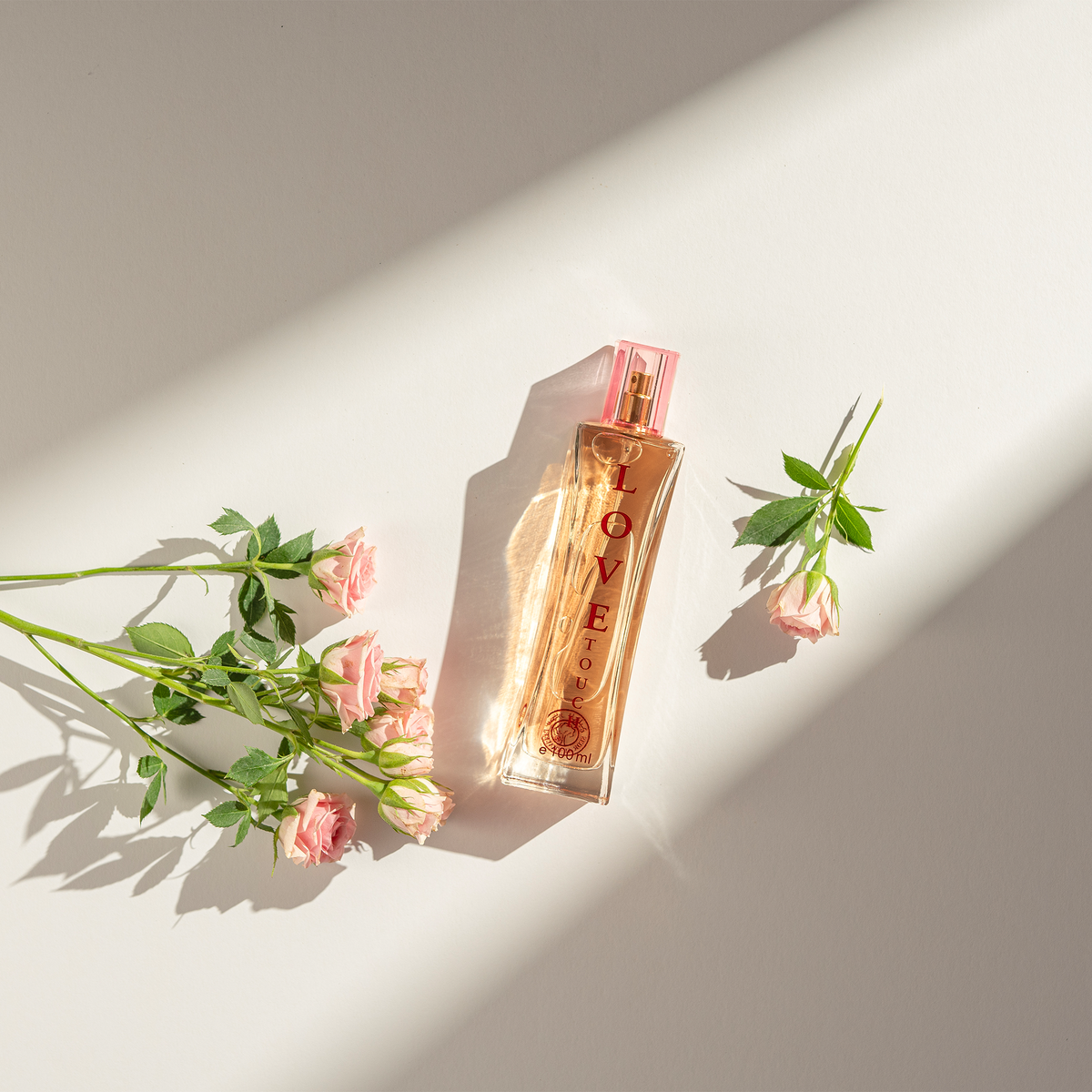 Love Touch Perfume 50 ml lying on a light surface between pink rose stems in sunlight