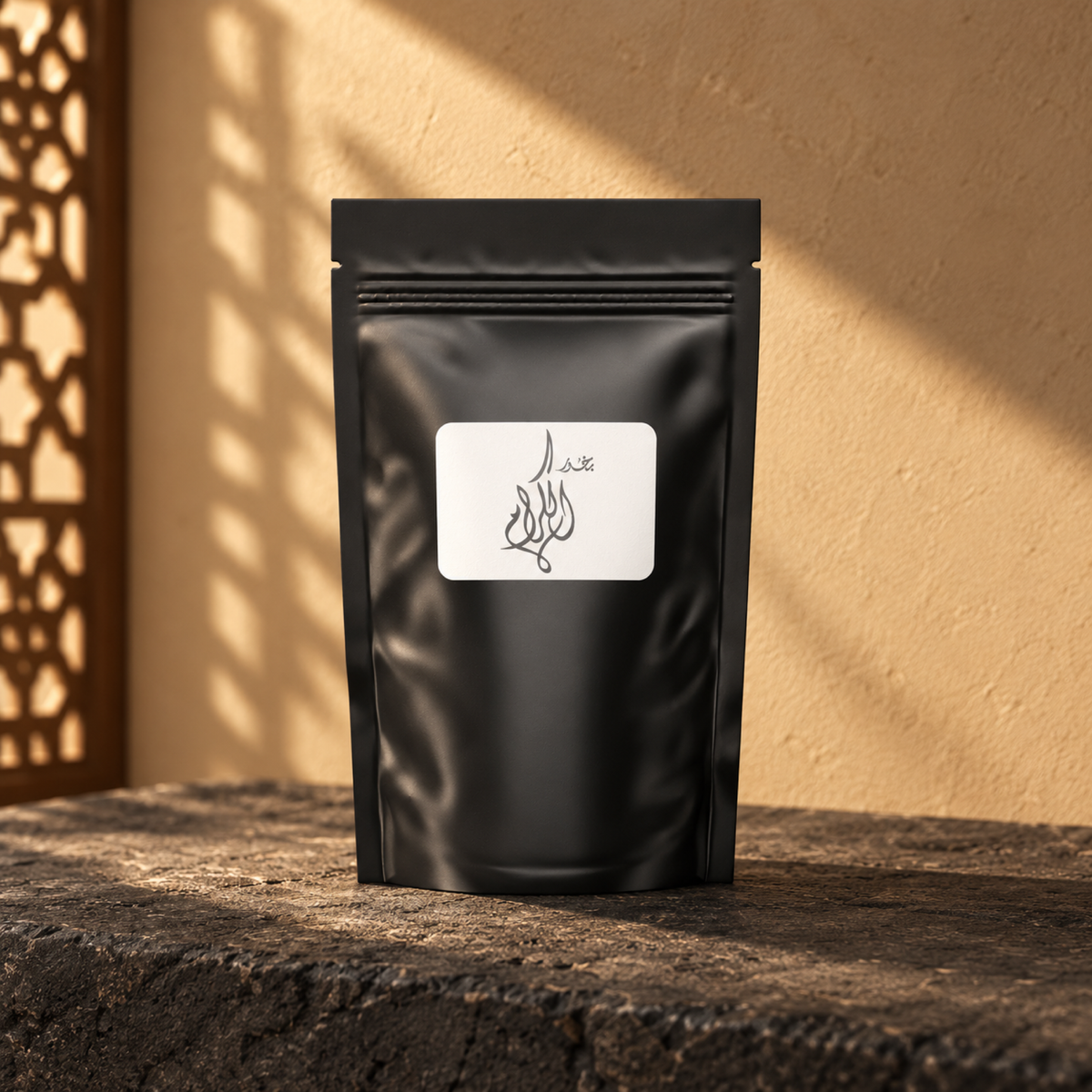 Al-Keram Bakhoor by Weight from Sedr Al-Khaleej; closed matte black pouch with a white label on dark basalt against a sand plaster wall, with angled sunlight and a wooden screen shadow.