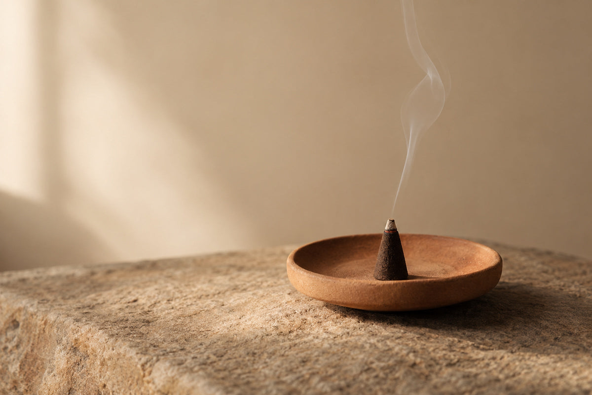 A lit incense cone giving off a thread of smoke, in a clay dish on a stone surface