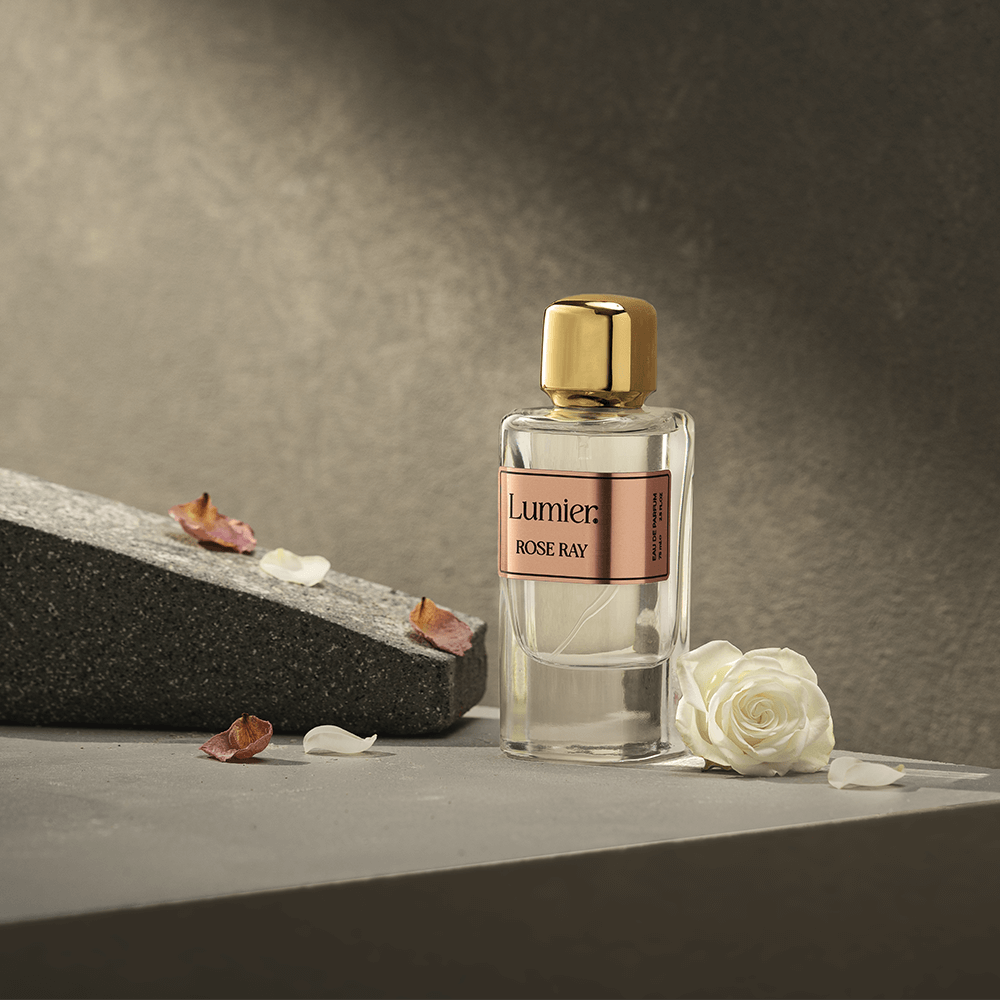 Rose Ray Perfume on a grey surface beside a slanted dark stone, a white rose and dried petals